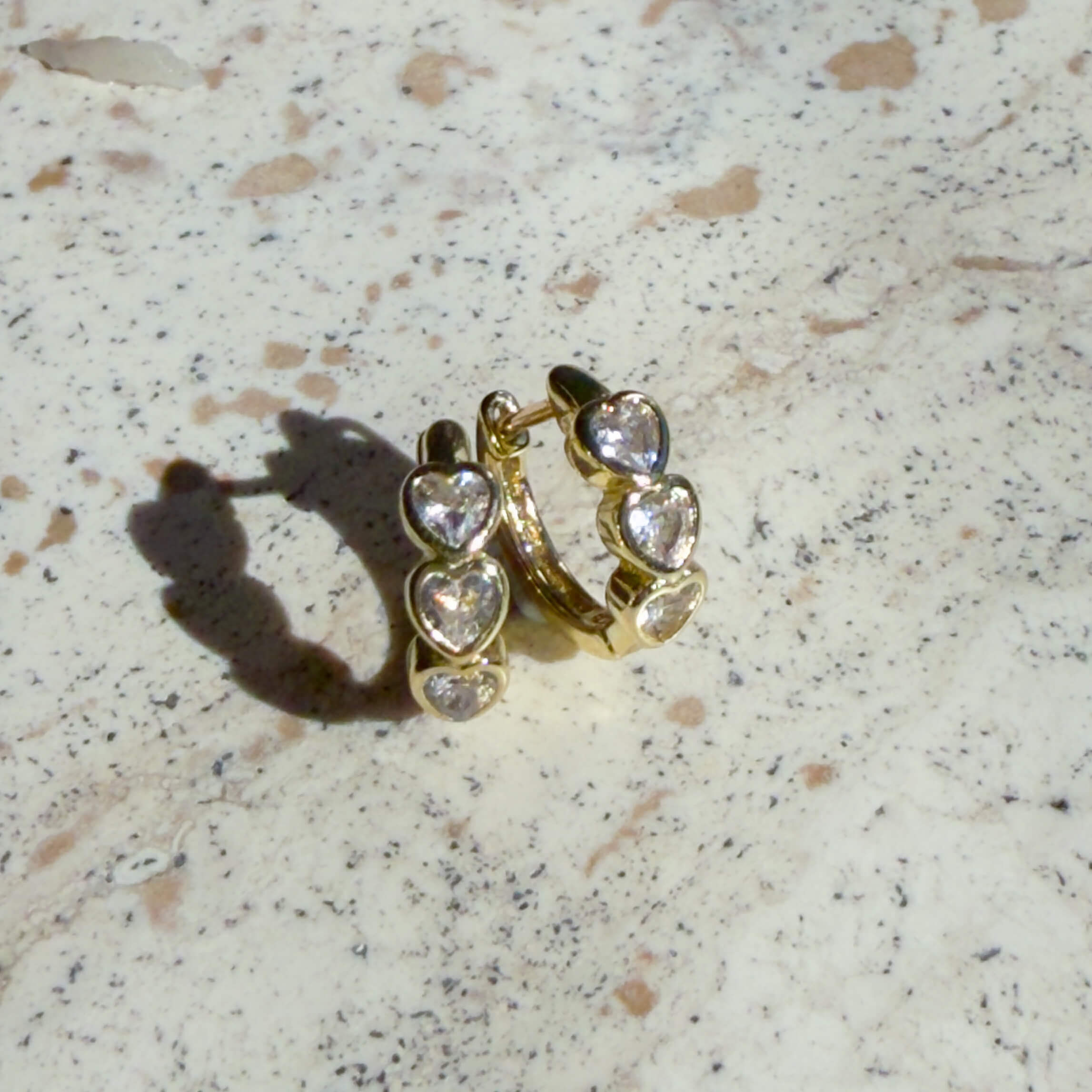 Gold hoop earrings with heart-shaped gemstones on a speckled surface